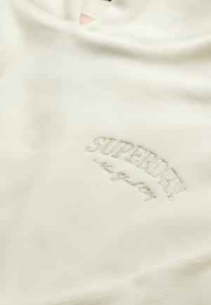 Tela de color crema con un sutil bordado del logo "SUPERDRY" y un texto en cursiva más pequeño debajo, todo sobre un material suave y texturizado.