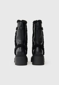 Botas de tornozelo em couro preto com um acabamento de pelo sintético felpudo, solas grossas e planas, e tiras ajustáveis na parte de trás para um encaixe seguro.