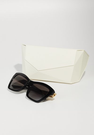 Lunettes de soleil noires surdimensionnées avec des verres dégradés, une monture en plastique épaisse et un accent doré sur la branche, accompagnées d'un étui géométrique blanc.