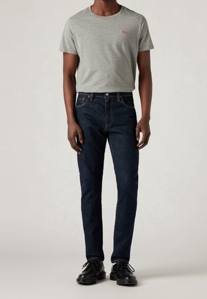 Person trägt ein graues T-Shirt mit kurzen Ärmeln, dunkelblaue Jeans und schwarze elegante Schuhe und steht vor einem schlichten hellen Hintergrund.