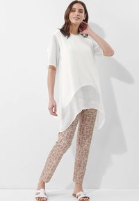 Witte doorschijnende top met korte mouwen boven een beige broek met geometrische patronen. De outfit wordt aangevuld met witte instapsandalen.