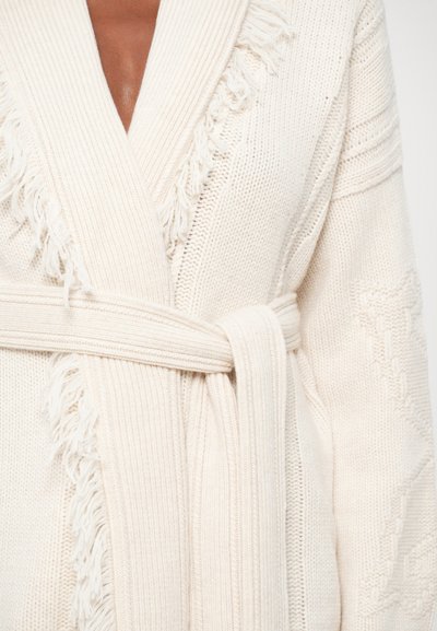 Cardigan tricotado em off-white com detalhes em franjas, textura canelada, gola larga e laço. Apresenta padrões decorativos nas mangas.