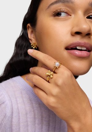 Mujer con pendientes dorados de estilo floral y dos anillos dorados, uno con perla, mirando de lado con la mano cerca del rostro, vestida con un suéter lavanda.