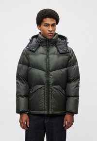 Tamno zelena podstavljena jakna s visokim ovratnikom, s crnom odvojivom kapuljačom i patentnim zatvaračem, naglašena bočnim džepovima i rebrastim manžetnama.