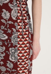 Robe bordeaux avec des motifs paisley et floraux, présentant un panneau géométrique avec des triangles rouges et crème. Détail de cordon de serrage à la taille.