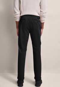 Osoba nosząca czarne eleganckie spodnie i czarne buty, stojąca tyłem do kamery na jednolitym beżowym tle.