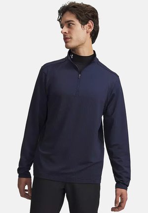 Young man wearing a navy blue quarter-zip pullover and black pants, looking to his left against a plain background.