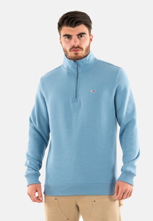 Sudadera de cremallera azul claro con cuello alto y puños acanalados. Presenta un pequeño logo en el pecho. Fabricada con un tejido suave y texturizado.