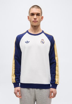 Mann som har på seg en hvit genser med lange ermer med blå ermer og gule striper, med Adidas- og Real Madrid-logoer.