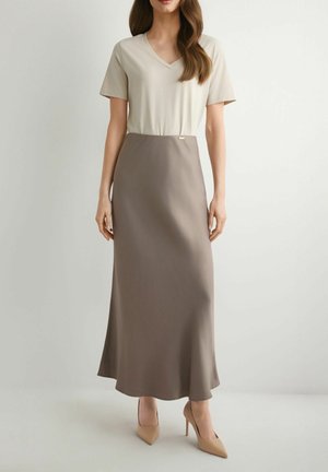 Kobieta w beżowym topie z krótkim rękawem i dekoltem w serek, brązowej spódnicy midi w kształcie litery A oraz beżowych szpilkach z noskiem w szpic, stojąca na białym tle.