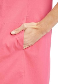 Abito in tessuto rosa con tasca laterale, texture morbida e design semplice. Mano appoggiata nella tasca, che evidenzia la funzionalità pratica.
