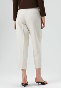 Crèmekleurige taps toelopende broek met een gladde textuur, voorzien van een mid-rise taille en subtiele details aan de achterzakken. Draagt zwarte sandalen met hak.