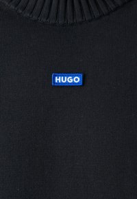 Pull en tricot côtelé noir avec une petite étiquette rectangulaire bleue placée au centre, affichant "HUGO" en lettres majuscules blanches.