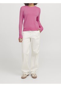 Pull rose côtelé à manches longues, associé à un pantalon large blanc et des baskets beiges, présentant une coupe décontractée et un design minimaliste.