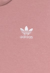 Sudadera de algodón rosa con un pequeño logotipo bordado en blanco de Adidas que presenta tres rayas y un diseño de trébol en el lado izquierdo del pecho.