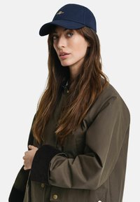 Marineblaue Mütze mit gebogenem Schirm und goldenem Logo, kombiniert mit einem braunen Mantel, der eine strukturierte Cord-Manschette und einen Knopfverschluss aufweist.