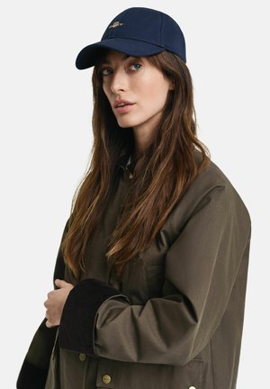 Marineblaue Mütze mit gebogenem Schirm und goldenem Logo, kombiniert mit einem braunen Mantel, der eine strukturierte Cord-Manschette und einen Knopfverschluss aufweist.