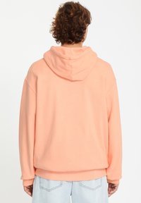 Haut à capuche couleur pêche avec cordon de serrage, au design oversize, avec des poignets côtelés et une texture lisse. Porté avec un jean bleu clair.