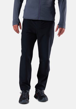 Mann, der schwarze Hose, schwarze und graue Wanderschuhe und eine dunkelgraue Zip-up-Jacke trägt, steht vor einem weißen Hintergrund.