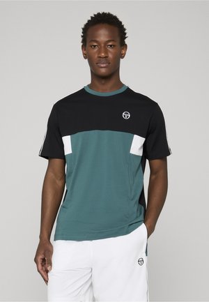 Giovane uomo che indossa una maglietta a blocchi di colore neri, turchesi e bianchi e pantaloncini bianchi con il logo di un marchio sportivo, in piedi davanti a uno sfondo grigio.