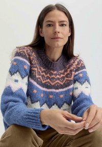 Pull en tricot dans des nuances de bleu, blanc et orange, avec un motif géométrique, un col rond et des poignets côtelés. Tissu doux et texturé.