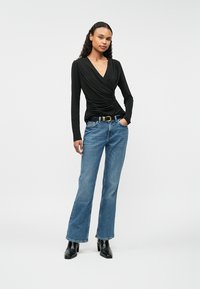 Sieviete ģērbusies melnā satinātā garā piedurkņu topā, zilās augstajā jostasvietā taisnās kājās džinsās ar melnu jostu un melnās potīšu zābakos, stāv ar roku kabatā.