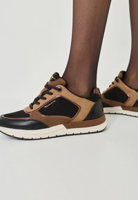 Braune und schwarze Sneaker mit einem strukturierten, gemischten Materialdesign. Sie verfügen über eine weiße Sohle und schwarze Schnürsenkel, die kontrastierende Paneele betonen.