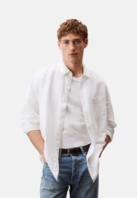 Camisa blanca de manga larga con cuello, botones en la parte delantera y un bolsillo en el pecho, superpuesta a una camiseta blanca, combinada con jeans azules.