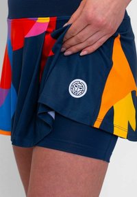 Falda atlética colorida con patrones abstractos en rojo, azul, naranja y amarillo; combinada con pantalones cortos ajustados de color marino y un detalle de logo redondo.
