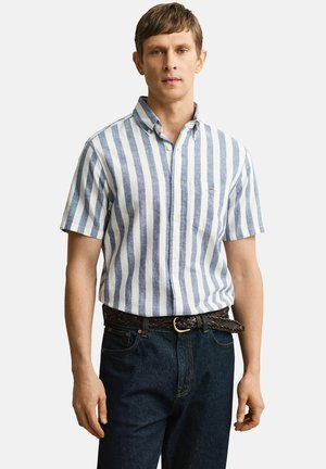 Mann trägt ein kurzärmliges Hemd mit blau-weißen vertikalen Streifen, das in eine dunkelblaue Jeans mit braunem geflochtenem Gürtel gesteckt ist.