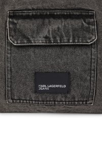 Primeiro plano de um bolso de denim preto texturizado com aba e etiqueta retangular preta com a inscrição "KARL LAGERFELD JEANS" em texto branco.
