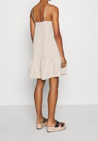 Kvinna iförd en beige ärmlös klänning med volangkant, i kombination med svarta plattformssandaler med dubbla spännen, stående mot en vit vägg.