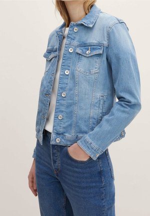 Kvinde iført lysblå denimjakke over hvid top med medium blå jeans, den ene hånd i lommen, foran ensartet baggrund.