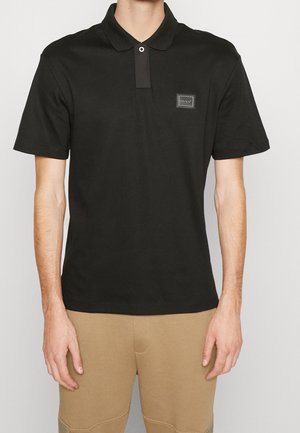 Homme portant un polo noir à manches courtes avec un petit écusson rectangulaire "Hugo" sur la poitrine et un pantalon beige, debout devant un fond uni.