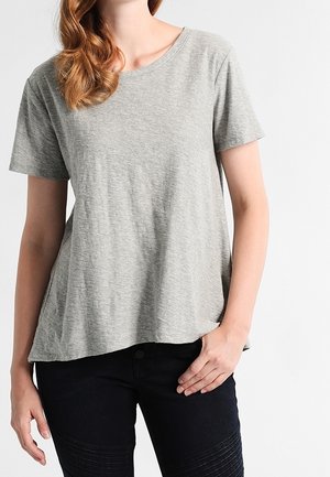 Kobieta ubrana w luźną, jasnoszarą koszulkę z krótkim rękawem i czarne spodnie, z jedną ręką opartą na biodrze.