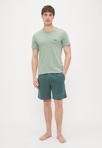 T-shirt de algodão verde claro com logótipo "BOSS" preto e calções verde-azulado. Ambos são casuais e de design simples. A modelo está descalça, em pé, com postura recta.