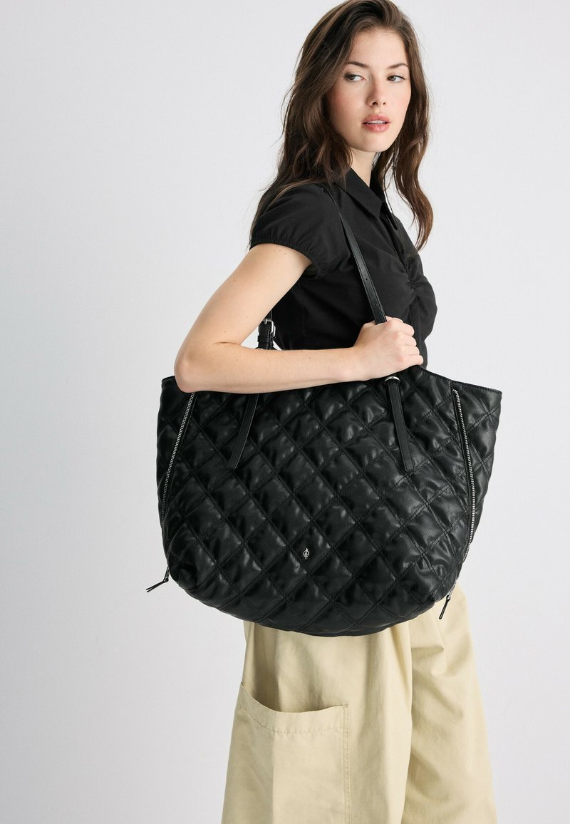 Jeune femme portant une chemise noire et un pantalon beige tient un grand sac à main noir matelassé sur son épaule sur un fond uni.