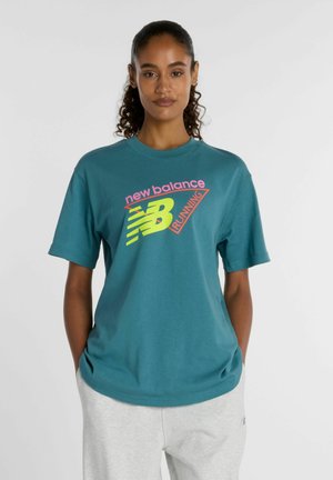 Frau mit lockigem Haar in türkisfarbenem New Balance Laufshirt und hellgrauer Hose steht mit den Händen in den Taschen vor neutralem Hintergrund.