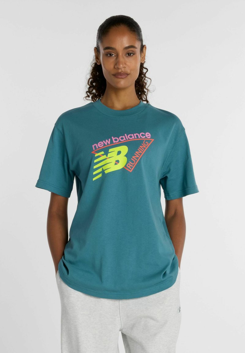 Femme aux cheveux bouclés portant un t-shirt de course New Balance bleu sarcelle et un pantalon gris clair, debout les mains dans les poches devant un fond uni.