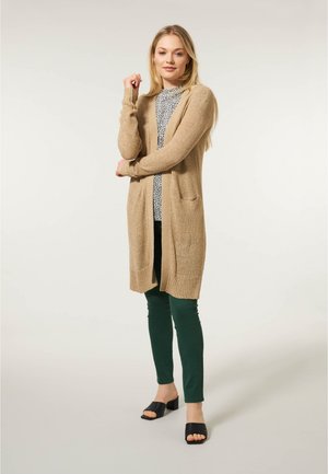 Beige gebreide cardigan met lange mouwen en open voorkant, met zakken en geribbelde zoom, gedragen over een bedrukt topje en groene broek.