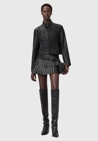 Μαύρο δερμάτινο στρατιωτικού στιλ μπουφάν με διακοσμητικά κουμπιά, συνδυασμένο με πτυχωτή μαύρη μίνι φούστα και μαύρες μπότες μέχρι το γόνατο.