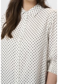 Femme portant un chemisier blanc boutonné avec des pois noirs, col pointu et manches froncées finissant aux coudes.