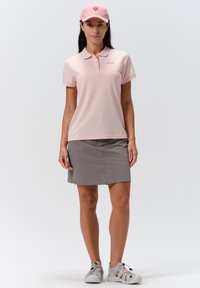 Vaaleanpunainen polo paita lyhyillä hihoilla ja kauluksella, yhdistettynä harmaaseen hameeseen. Mallilla on päällään vaaleanpunainen lippalakki ja harmaat kengät.