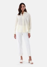 Camicia button-up giallo chiaro con un motivo a pois, abbinata a pantaloni bianchi con risvolto e sandali beige. Tessuto morbido, vestibilità rilassata.