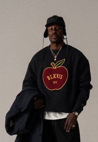 Sweatshirt preta com um gráfico de maçã vermelha com detalhes amarelos e texto "BLKVIS NY". Modelo usa um chapéu tipo bucket preto e segura um casaco.