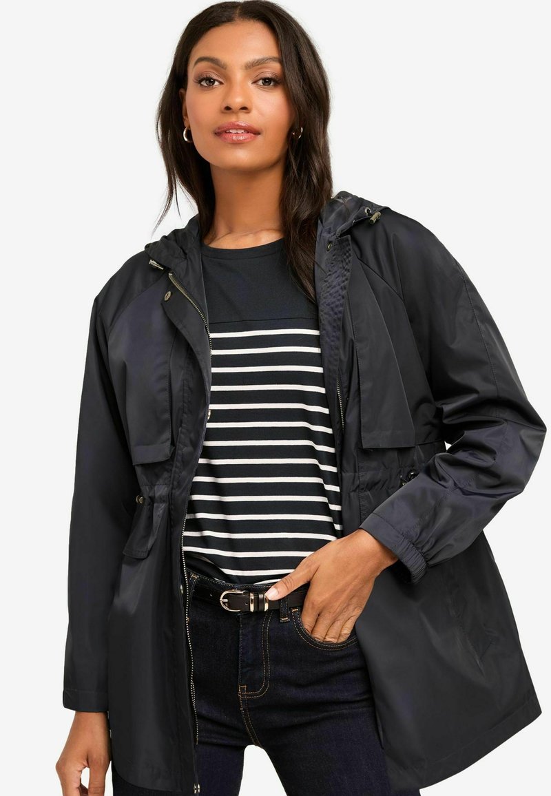 Marineblauwe waterdichte jas met capuchon, voorzien van een rits aan de voorkant, twee vo zakken en elastische manchetten, gelaagd over een zwart-wit gestreept shirt.
