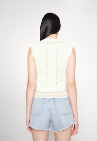 Valkoinen neulottu ilman hihoja oleva toppi, jossa on teksturoitu kuvio ja ribbireunat, yhdistettynä vaaleansinisiin farkkushortseihin.