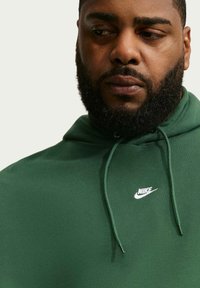 Άνδρας με γένια που φοράει πράσινο φούτερ Nike, κοιτάζοντας προς τα κάτω και ελαφρώς στο πλάι, με φόντο ένα απλό ανοιχτό χρώμα.