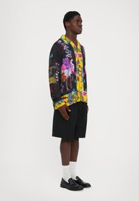 Bărbat îmbrăcat cu o cămașă cu imprimeu floral și cu păsări, cu mâneci lungi, pantaloni scurți negri, șosete albe și mocasini negri, stând în fața unui fundal alb simplu.