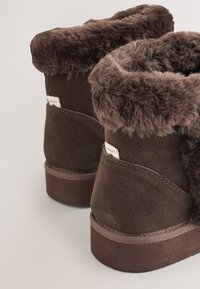 Hnědé kotníkové boty s měkkým, chlupatým kožíškem a semišovým povrchem. Mají silnou podrážku a prošité detaily na okrajích.